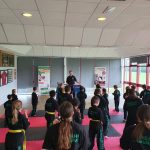 A class of children in black martial arts uniforms in the guard position face their instructor