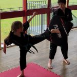 A girl in a martial arts uniform does a sidekick into a kickshield with a partner